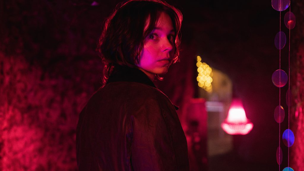 "Person standing in a dimly lit tunnel with exposed stone walls, illuminated by vibrant pink, purple, and blue lights and surrounded by hanging colorful circles, creating a mysterious and moody atmosphere."